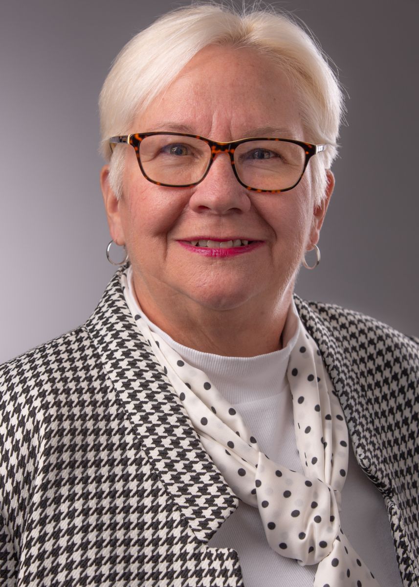 Ms. Sherrie Rowe, a Caucasian female with short white hair and glasses, wears medium silver hoop earrings, a white turtle neck with a black and white polk-a-dot scarf, and a black and white dress jacket with a square pattern weave.