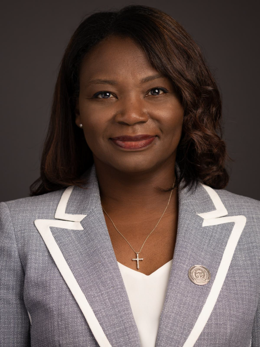 Tara Jenkins, an African American female with shoulder length brown hair, smiles wearing a light grey suit jacket with white trim and a white v-neck dress shirt against a dark grey background.